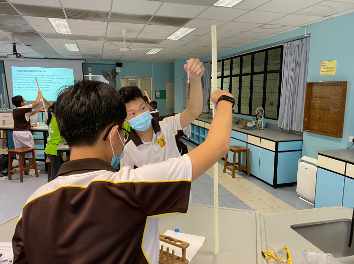 Two masked students in school uniforms conduct a science experiment with a vertical tube in a laboratory classroom.